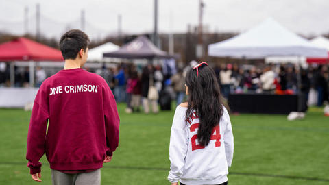 Two students with their back to the camera in Harvard sweatshirts walking towards tents during the Harvard-Yale game.