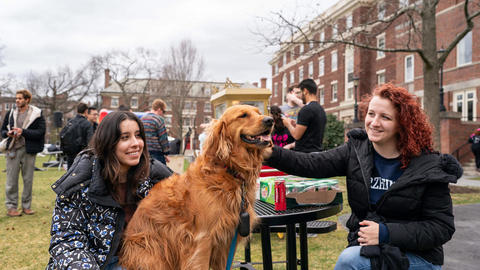 Two students pose for a photo with a proctor's dog on the Quad lawn.