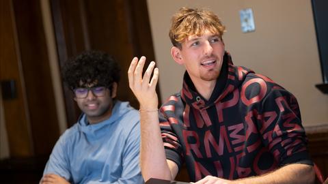 Two students depicted in a classroom with the student int he foreground engaged in dialogue.