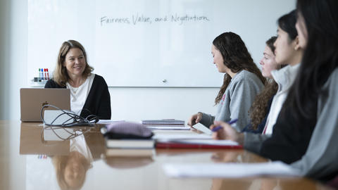 A professor sits at the head of a table during a course with four students appearing on her left side.