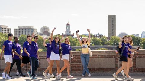 Students in purple and yellow Yard shirts cross the bridge over the Charles River as they wave to the camera.