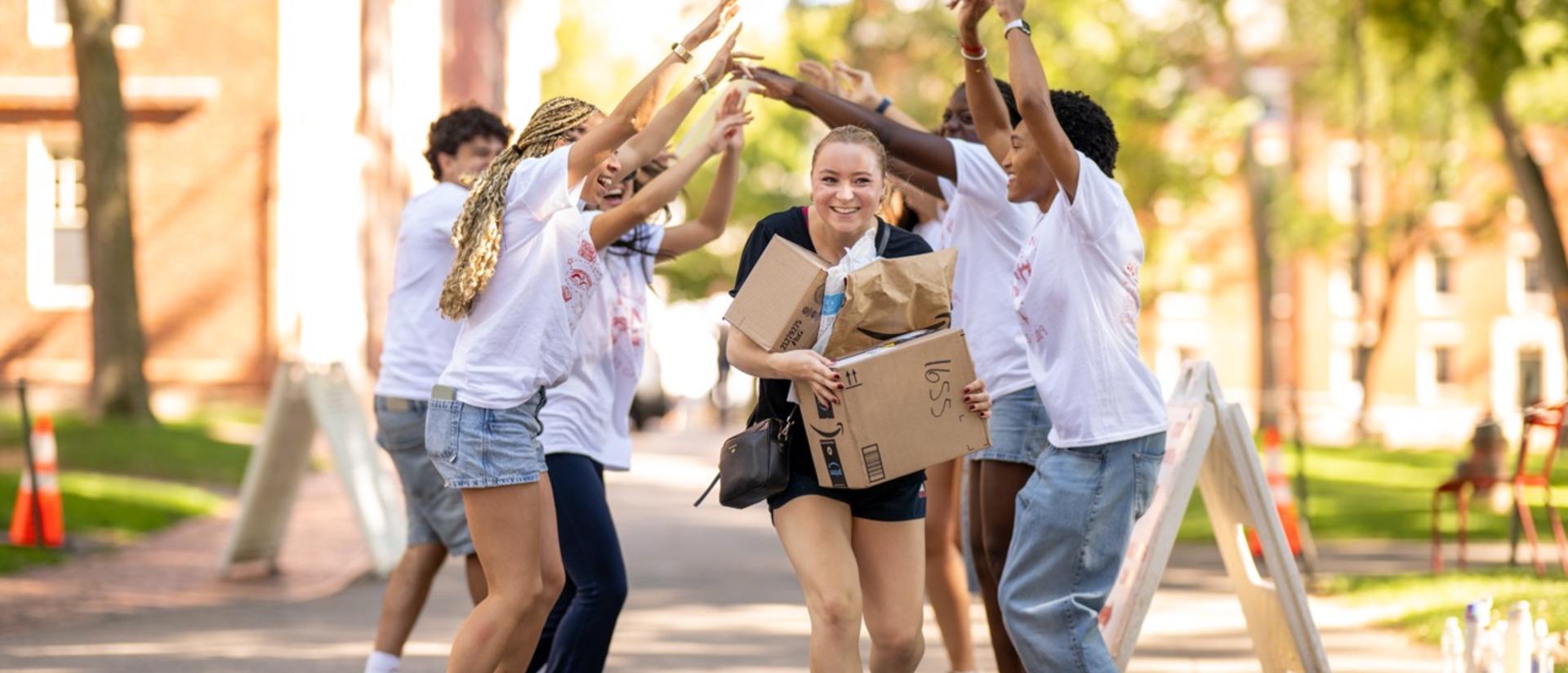 First-year student with a stack of packages from the mail center goes through a tunnel of student orientation works making a bridge with their hands.