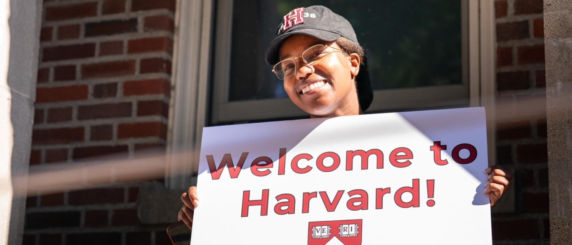 A student holding a "Welcome to Harvard" sign outside of a first-year dorm during move-in.